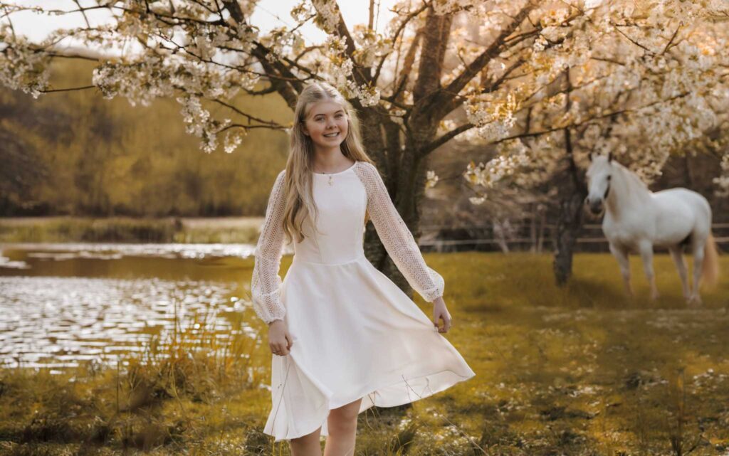 Fotograf Sarah-Simone tilbyder børnefotografering i Slagelse, Korsør og resten af Vestsjælland En konfirmand i en hvid kjole står smilende under et blomstrende træ ved en sø, med en hvid hest i baggrunden